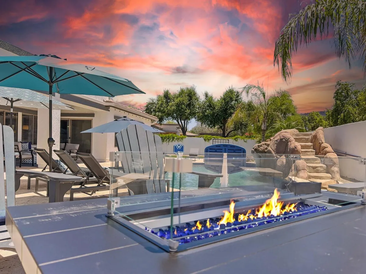 A beautifully landscaped backyard featuring a sparkling swimming pool, a cozy fire pit surrounded by lounge chairs, shaded areas with umbrellas, and lush greenery. The scene is tranquil, inviting relaxation, socializing, and outdoor dining with a view of the pool and sunset-ready atmosphere.