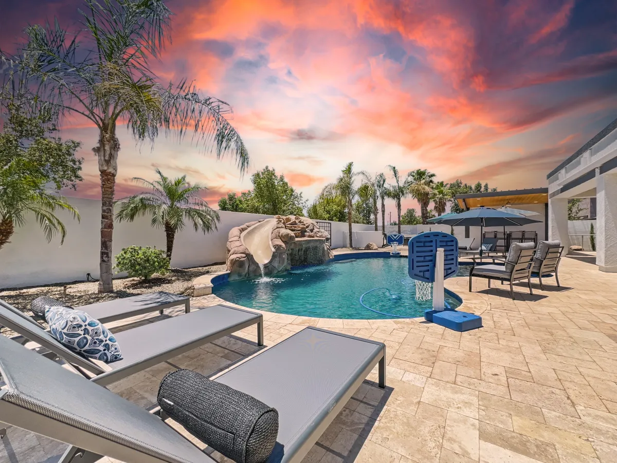 A lively backyard scene featuring a sparkling swimming pool with a waterslide, surrounded by lounge chairs and palm trees. There is an outdoor dining area visible, perfect for meals poolside. The setting is joyful and inviting, suggesting a space where families and friends gather for memorable summer barbecues and pool parties.