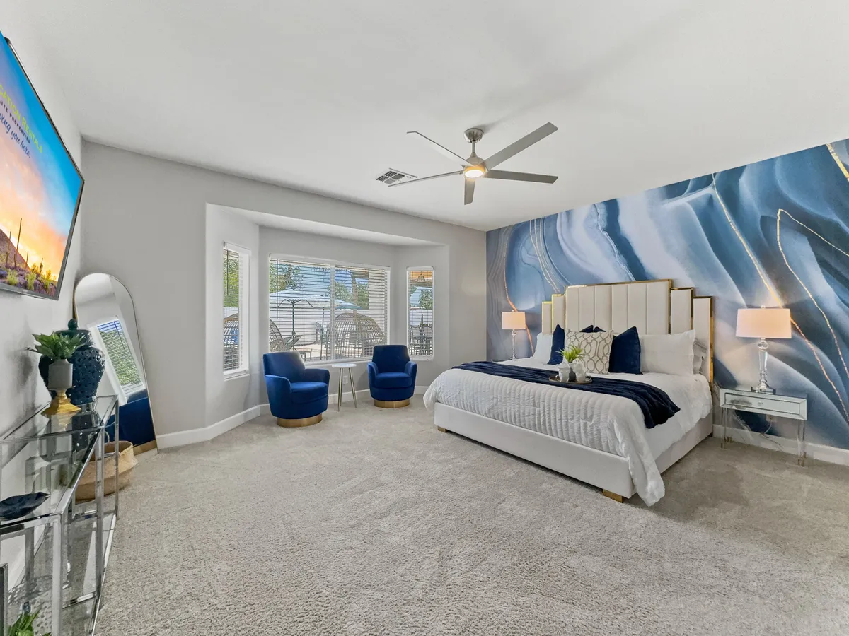 A serene king bedroom with a spacious king-size bed adorned with plush bedding, large abstract wall art above the headboard, a comfortable seating area with blue chairs, and a modern ceiling fan overhead. Natural light filters in, creating a calm and luxurious atmosphere.