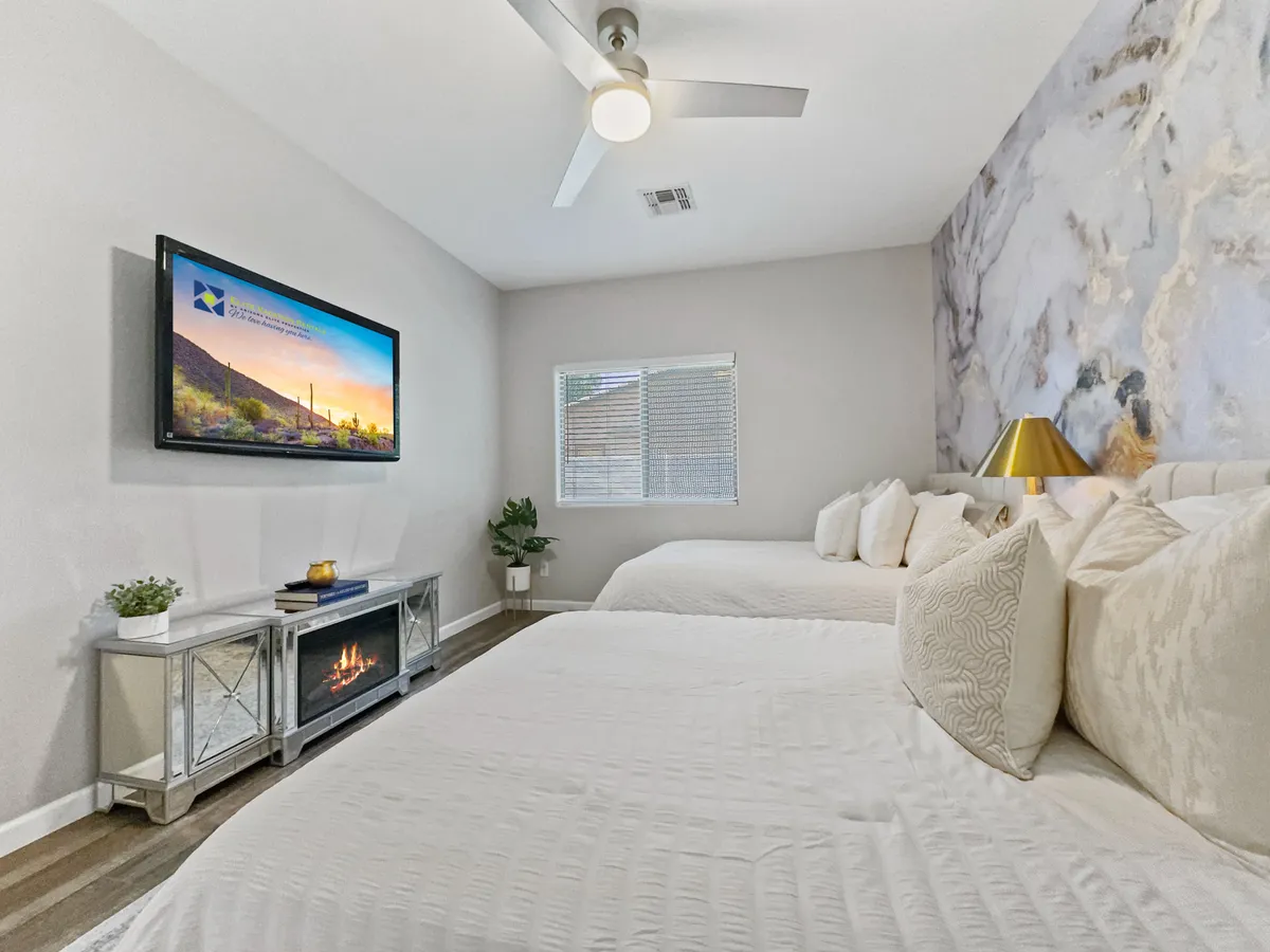 A modern bedroom featuring two beds with crisp white bedding, stylish accent pillows, a mounted TV, a sleek mirrored console table with a fireplace feature, and touches of greenery. The room is bright and spacious, perfect for families or friends.