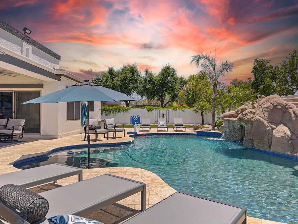 A tranquil backyard scene featuring a sparkling swimming pool, lounge chairs under umbrellas, and a natural rock formation. The setting invites swimming, sunbathing, and outdoor dining, with a spacious layout perfect for gatherings.