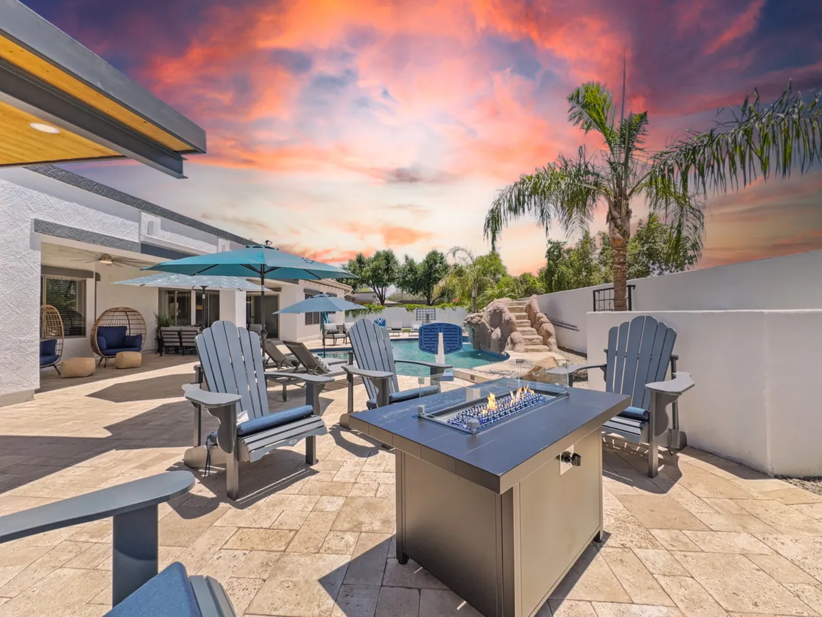 A beautiful evening scene showcasing a fire pit surrounded by lounge chairs, with the pool and umbrellas in the background. Lush landscaping and open views create a tranquil yet joyful setting, perfect for gathering around the fire under the Arizona sky.