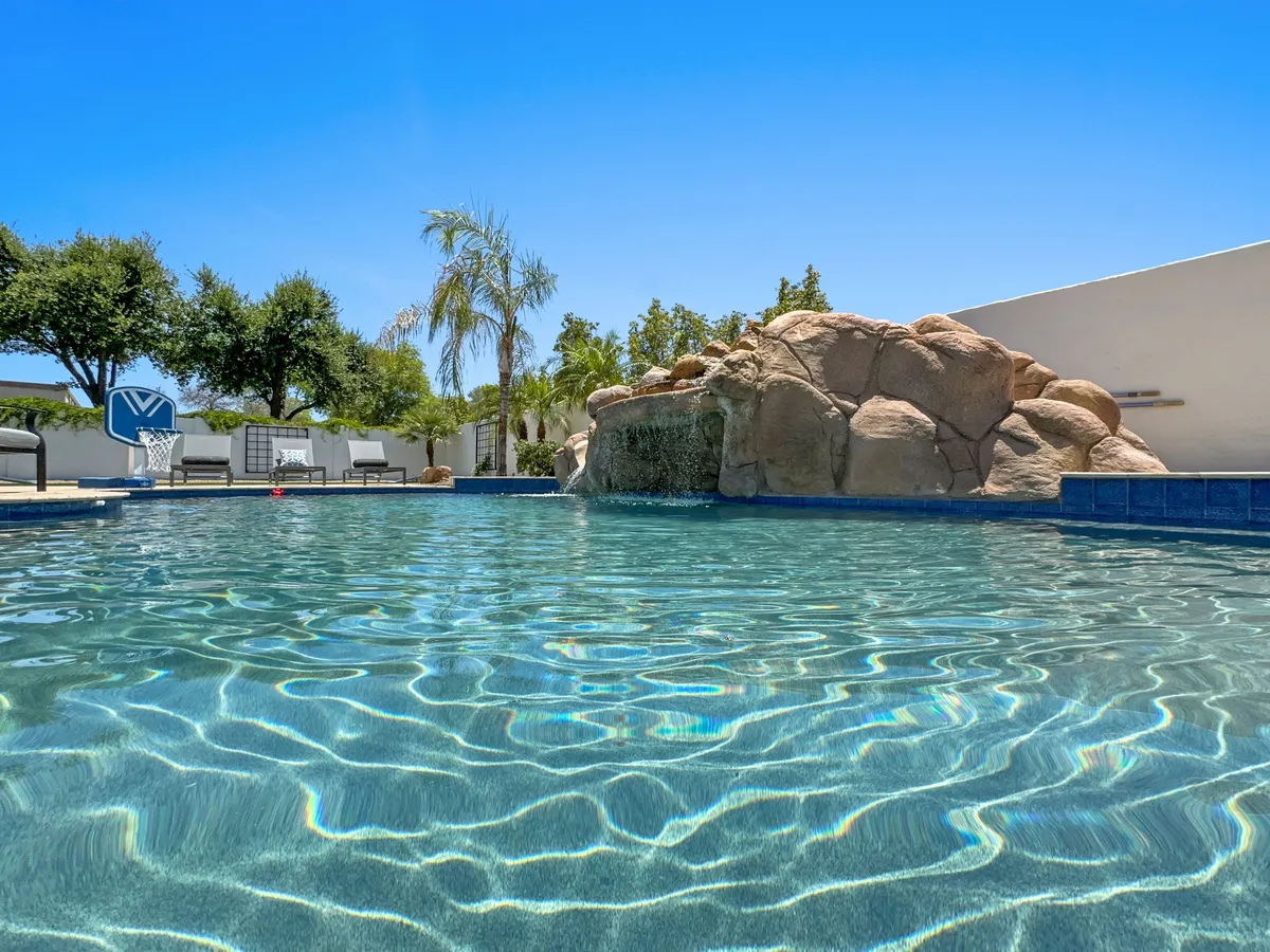 A wide exterior view displaying the swimming pool, rock formation, waterfall, palm trees, and multiple seating areas. The space is designed for swimming, sunbathing, and both lively and tranquil gatherings, highlighting the home’s resort-like feel.