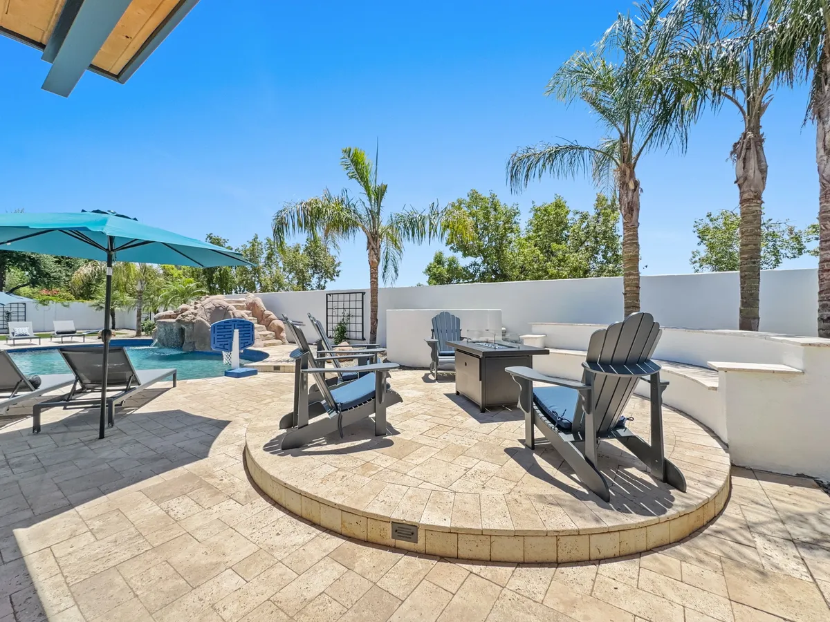 A beautifully landscaped backyard featuring a sparkling pool surrounded by lounge chairs, umbrellas, a cozy fire pit, and swaying palm trees. The scene is set for both relaxation and lively gatherings, with inviting seating areas and a tranquil pool reflecting the sky.