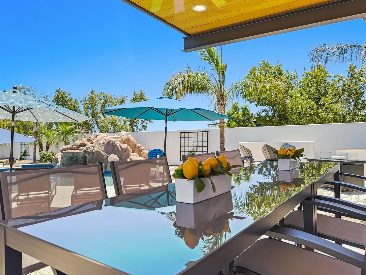 An outdoor dining table set under umbrellas, with a lemon centerpiece, overlooks the pool area. The scene is bright and cheerful, ideal for al fresco meals, pool parties, or enjoying a quiet morning coffee with a view of the landscaped grounds.