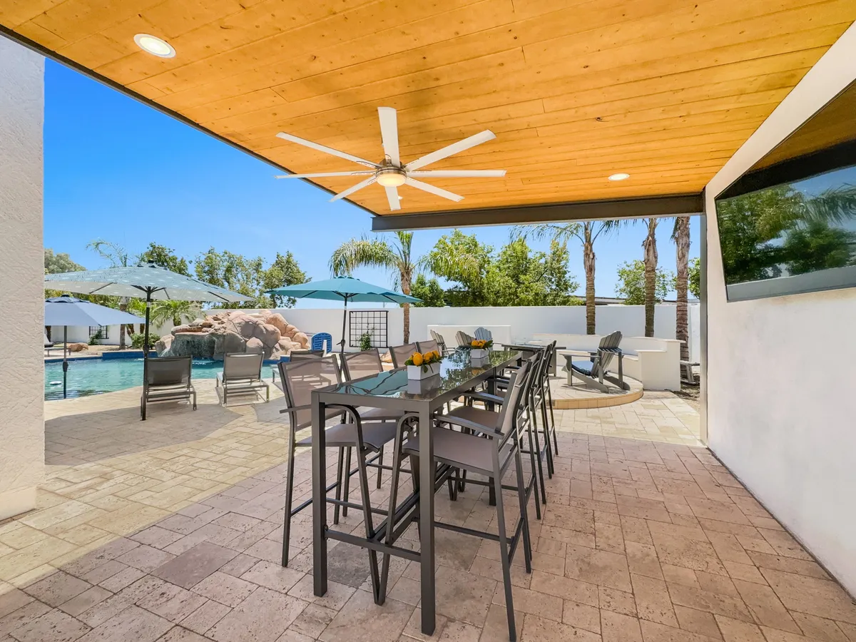 A vibrant outdoor living space with a large dining table set beside the swimming pool, shaded by umbrellas and surrounded by palm trees. There is also a ceiling fan above, and a TV visible for outdoor entertainment, all suggesting a sociable atmosphere for al fresco meals, parties, and relaxation.