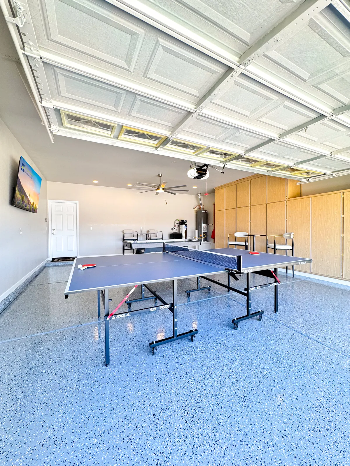 An energetic game room setup with a ping pong table, garage-style door, TV screen, and storage cabinets, creating an inviting atmosphere for family tournaments and friendly competitions.