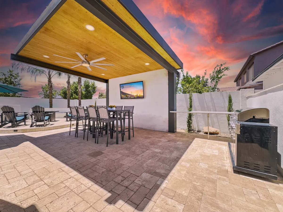 A cozy outdoor space with an inviting dining table, built-in BBQ grill, outdoor television, and lush landscaping. There's a ceiling fan overhead and landscape plants enhancing the relaxing ambiance. The scene is designed for alfresco meals, grilling, and watching big games outdoors.