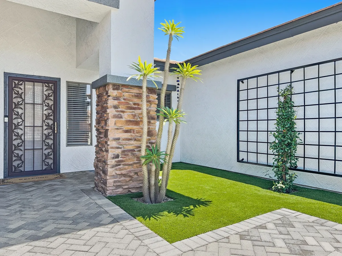A welcoming exterior entrance with a decorative door, potted plants, a stone wall, and a lush grassy area. The scene is calm and inviting, representing the very first impression of the property.