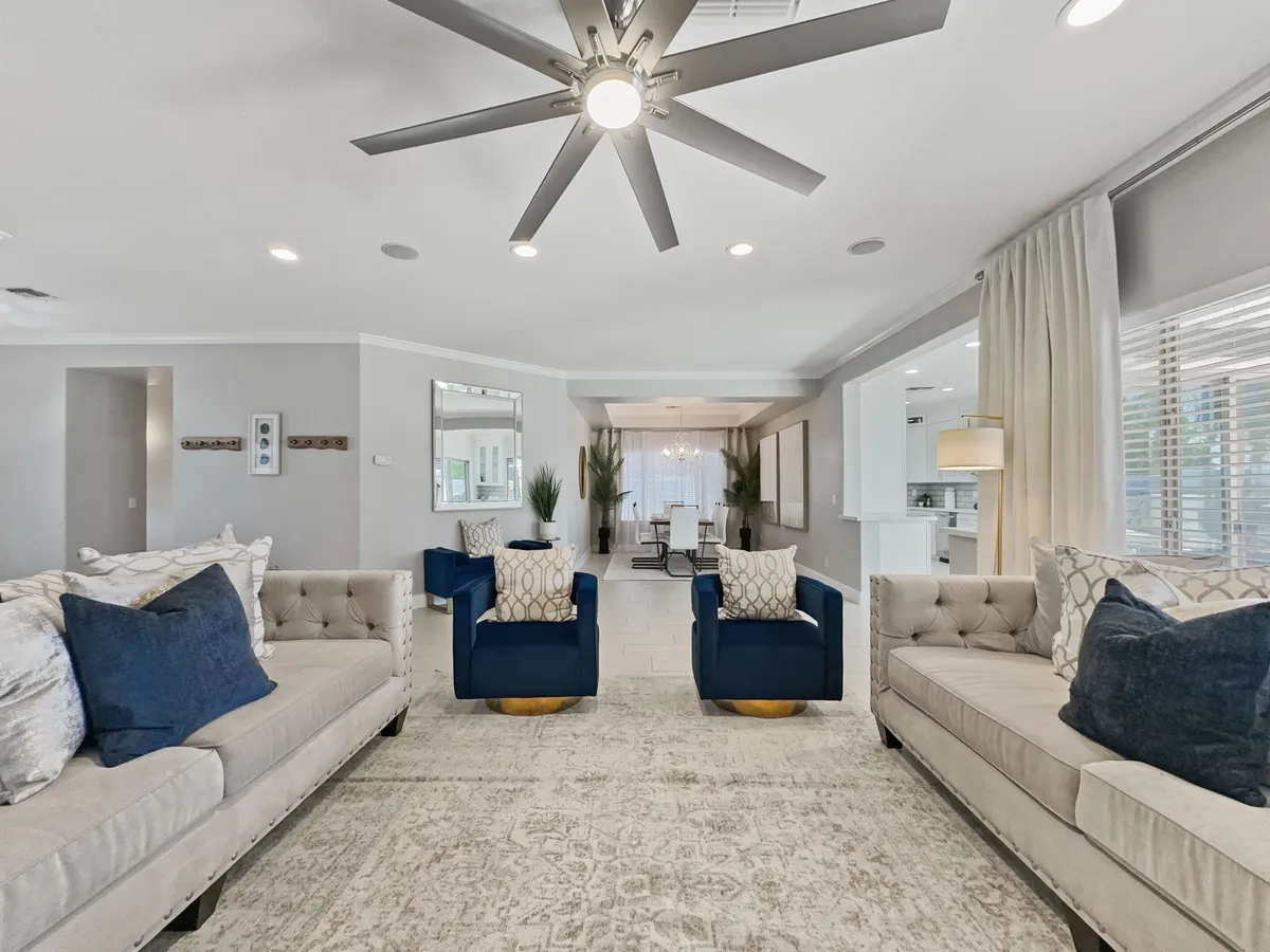 This image features a welcoming open-plan living area with a plush sofa set, blue accent chairs, a ceiling fan, and an expansive dining area visible in the background. The room is bathed in natural light, creating a bright and comfortable atmosphere perfectly suited for gatherings and family time.